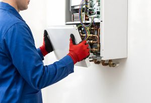 Technician removing the cover from a wall-mounted boiler with exposed pipes and wiring