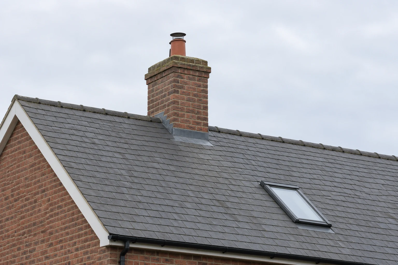 Brick chimney above a slate roof ridge, with a roof window lower down the slope.